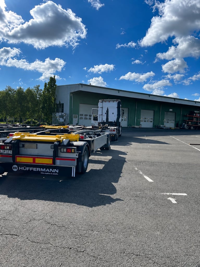 Huffermann flatbed trailers parked in front of industrial warehouse building under blue sky with white clouds